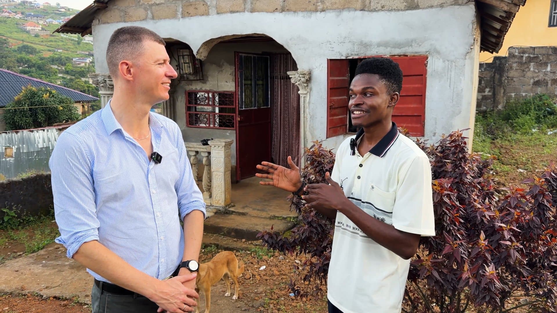 Patrick gesturing as he explains daily life to Ladislas Maurice in front of his house, with a dog in the yard and unfinished buildings on the hillside behind.