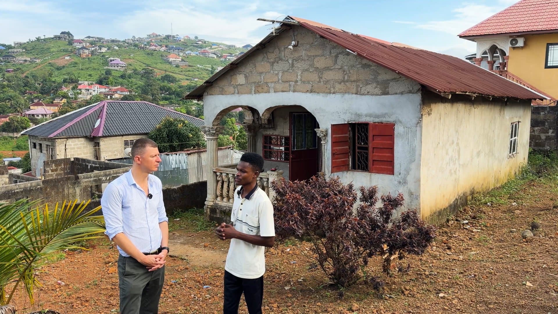 Ladislas Maurice talking with Patrick outside his single-story house in the Freetown hills, with the city spread out behind them.