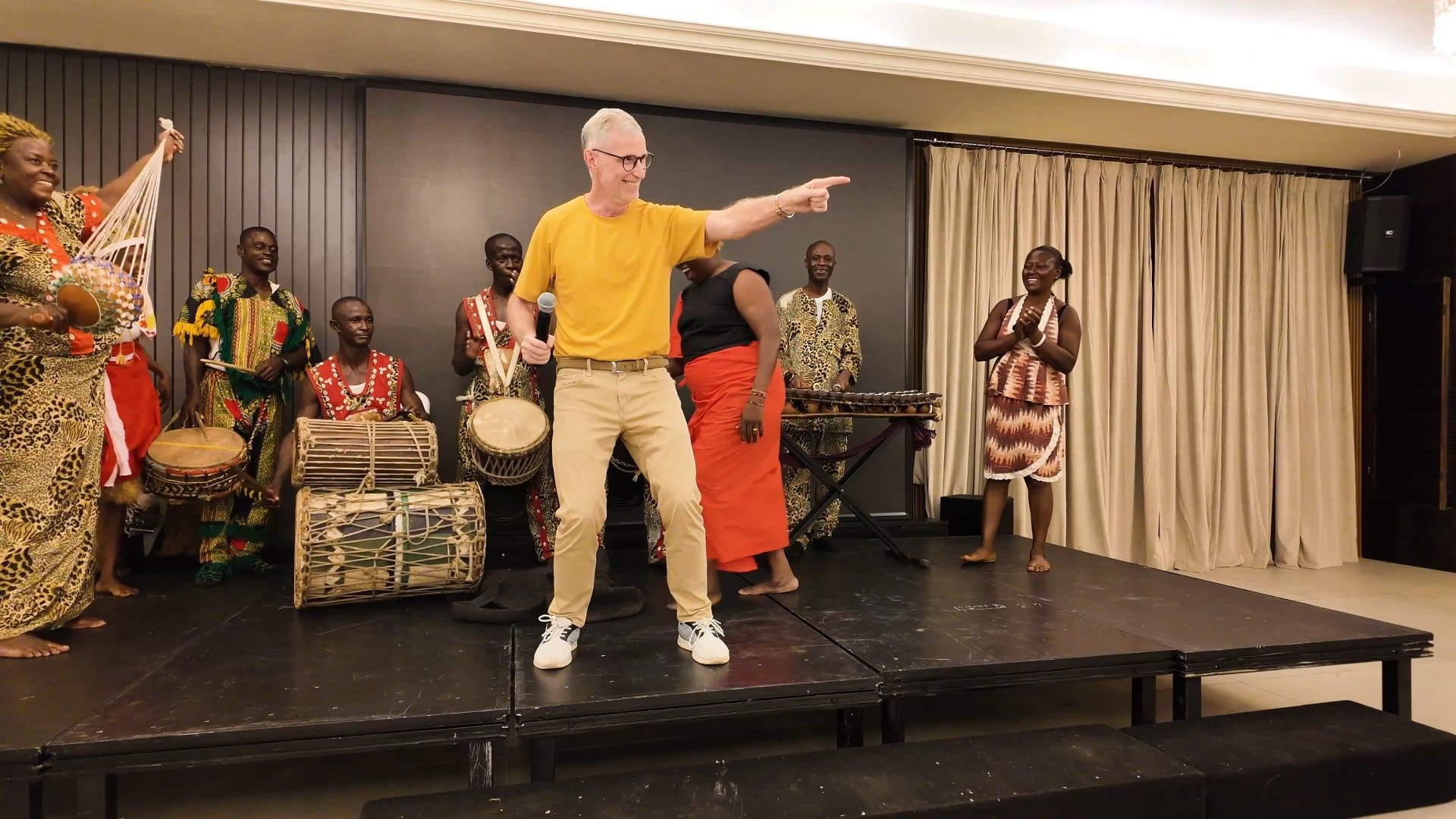 Stephen Barnes dancing on stage with a microphone, surrounded by traditional drummers and dancers at the delegation reception.