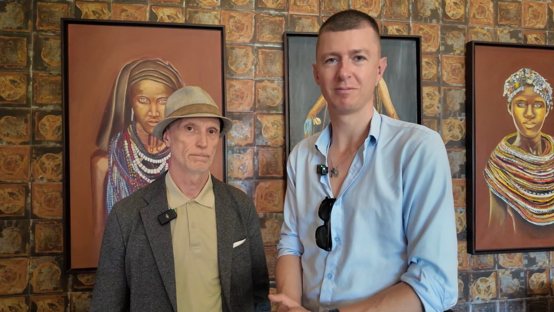 Stephen Barnes, in a grey hat and blazer, standing with Ladislas Maurice in front of portraits of Sierra Leonean women.