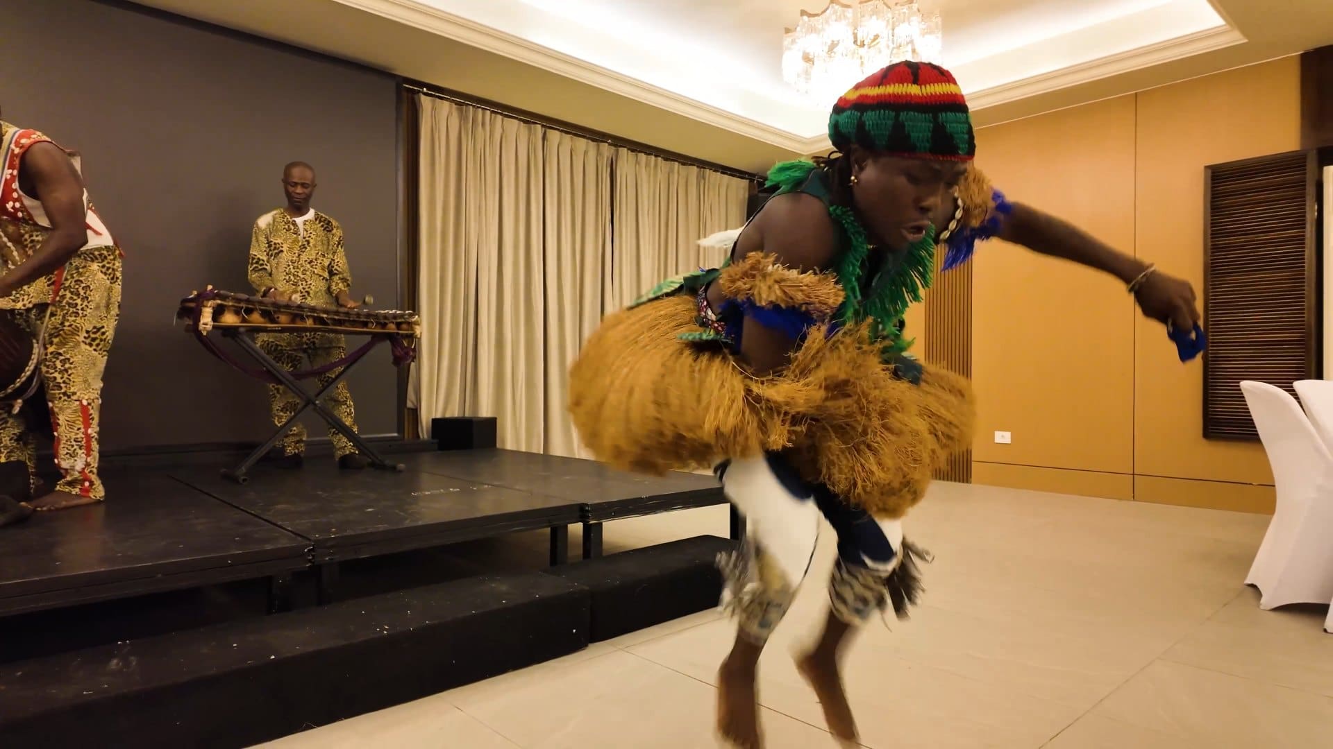 A dancer in a raffia costume and a red, green, and yellow knit hat mid-jump during a traditional performance in Freetown.